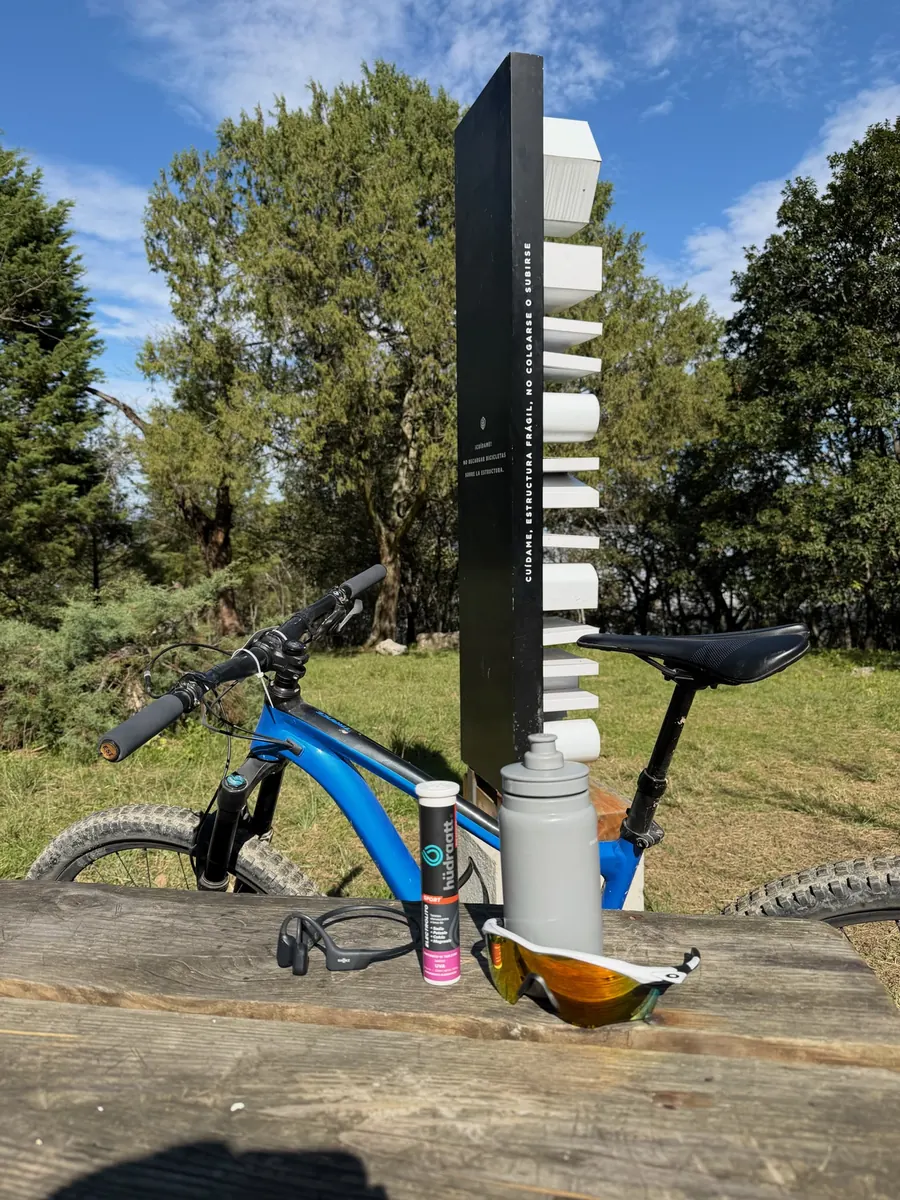 Tubo, botella y gafas sobre una mesa de madera frente a una bicicleta de montaña