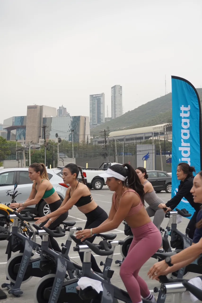 Grupo de mujeres en bicicletas de spinning al aire libre junto a una bandera de Hudraatt