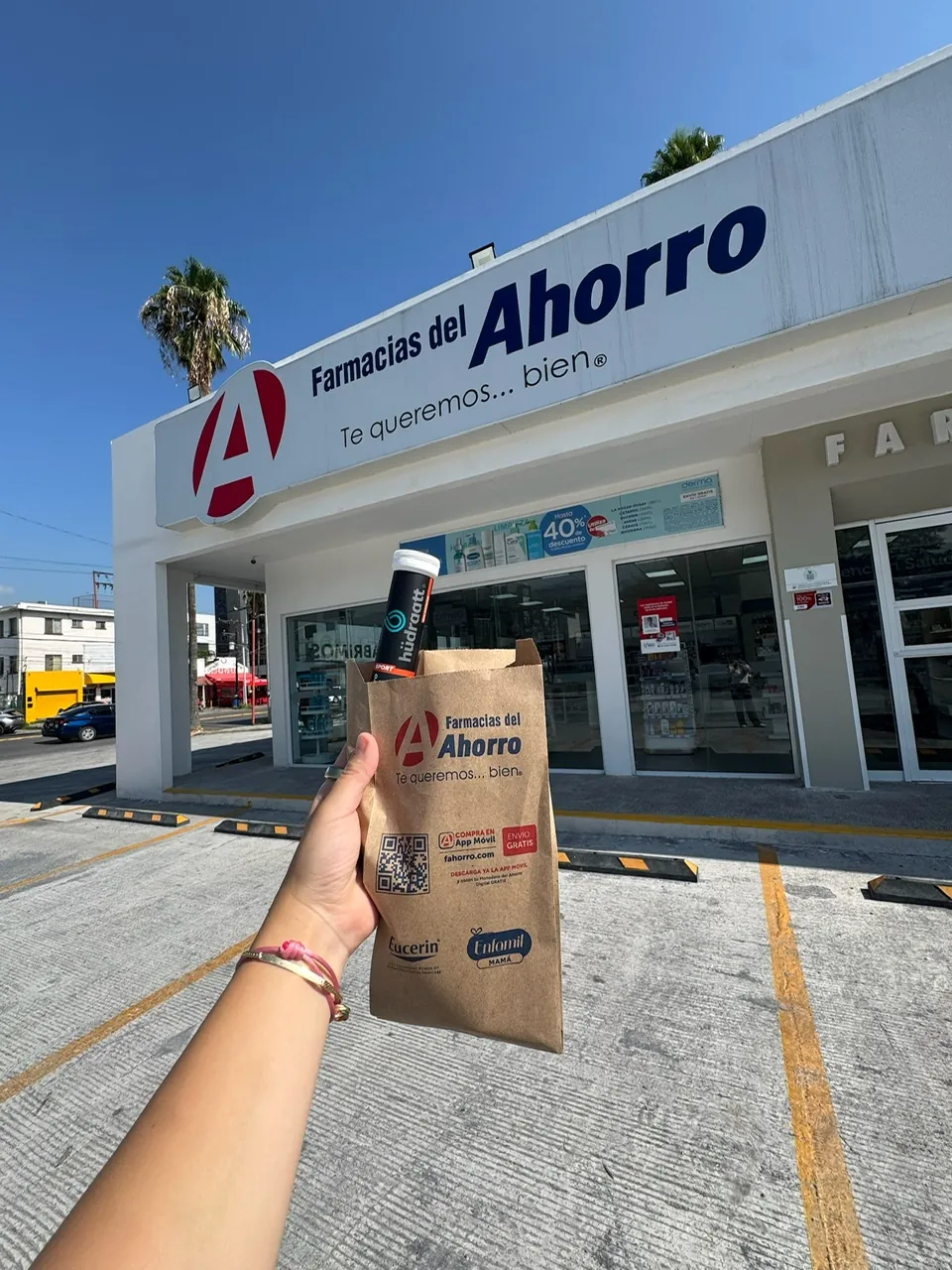 Mano con un tubo de Hudraatt y una bolsa frente a una sucursal de Farmacias del Ahorro