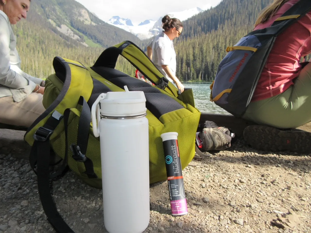 Tubo de Hudraatt Uva y una botella blanca junto a una mochila verde frente a un lago de montaña