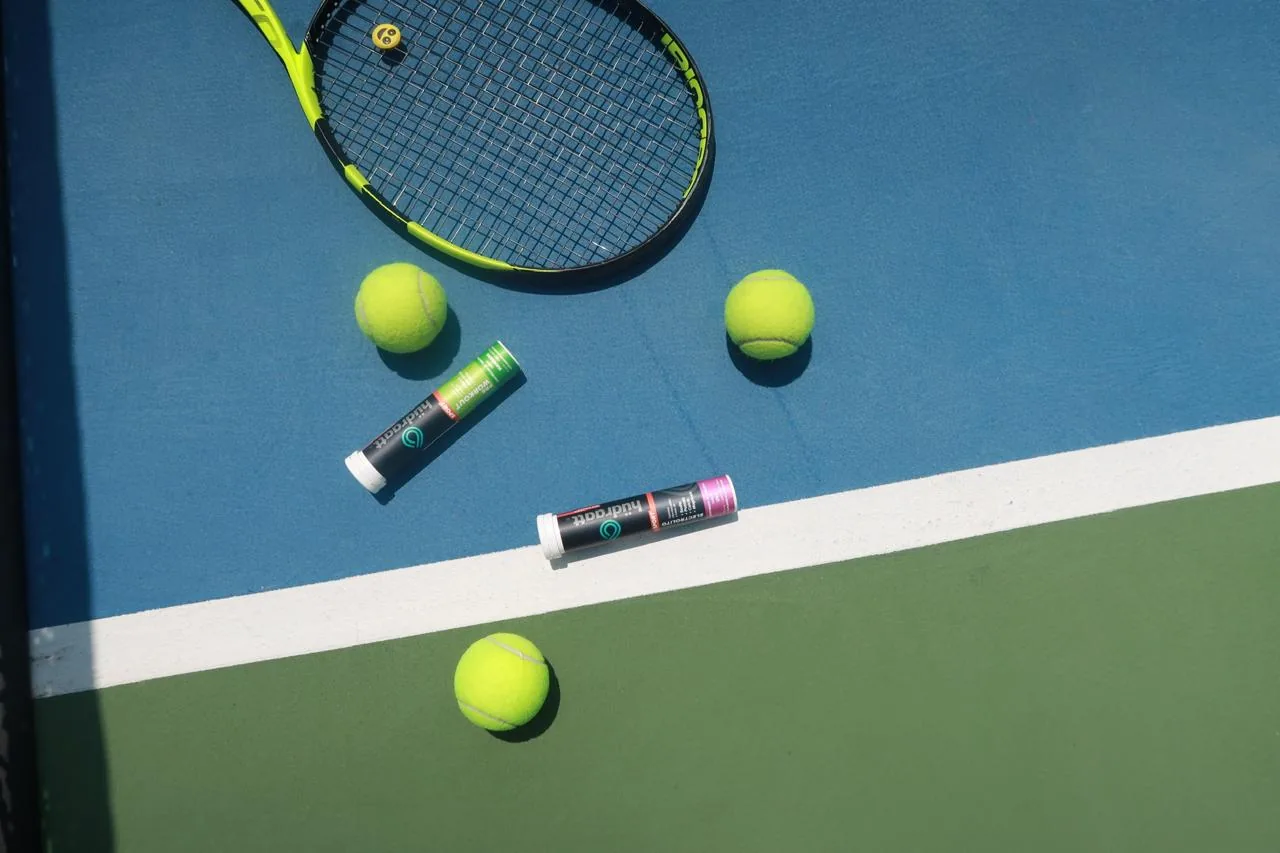 Tubos de Hudraatt Uva y Manzana sobre una cancha de tenis, junto a una raqueta y pelotas